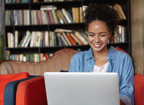 Happy pretty dark-skinned female in jean shirt sitting at sofa using laptop, typing something and browsing internet while being in reading hall or library.