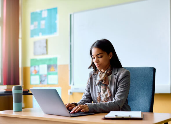 Hispanic teacher working on her laptop in classroom at school.