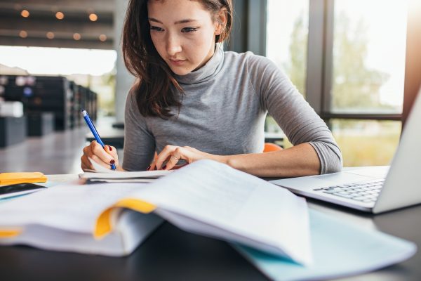 Close up image of a young female student doing assignments in library. Asian woman taking notes from textbook.